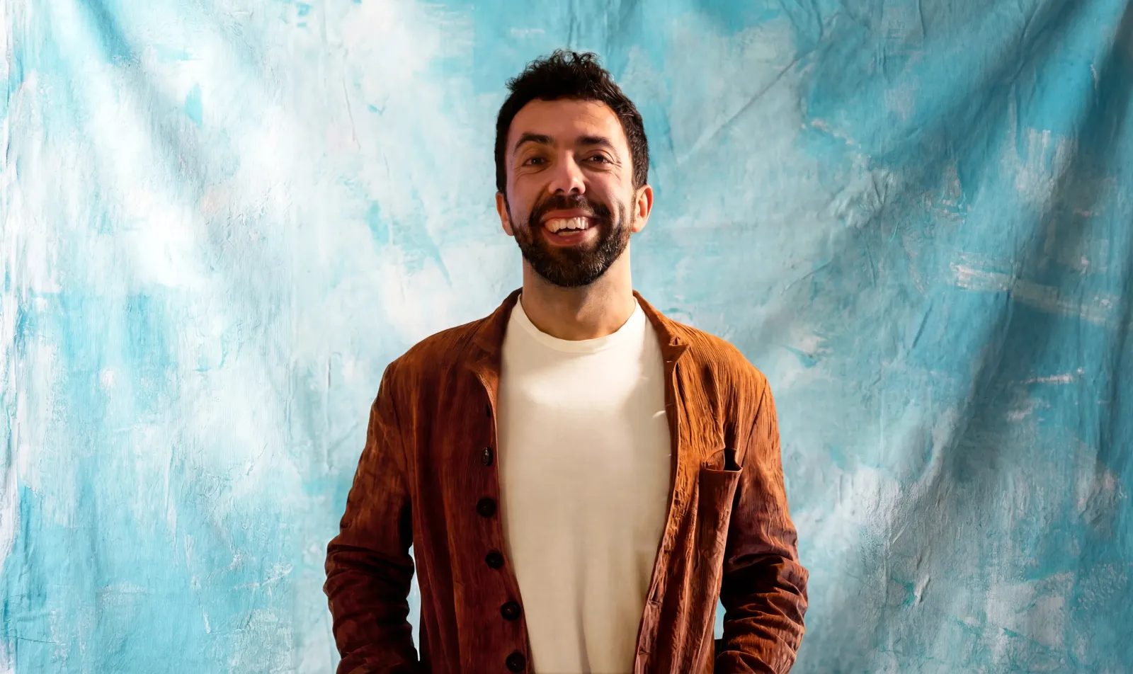Edoardo L'Astorina, CEO / Founder of Blu Frame, smiling against an artistic turquoise backdrop in a rust jacket.