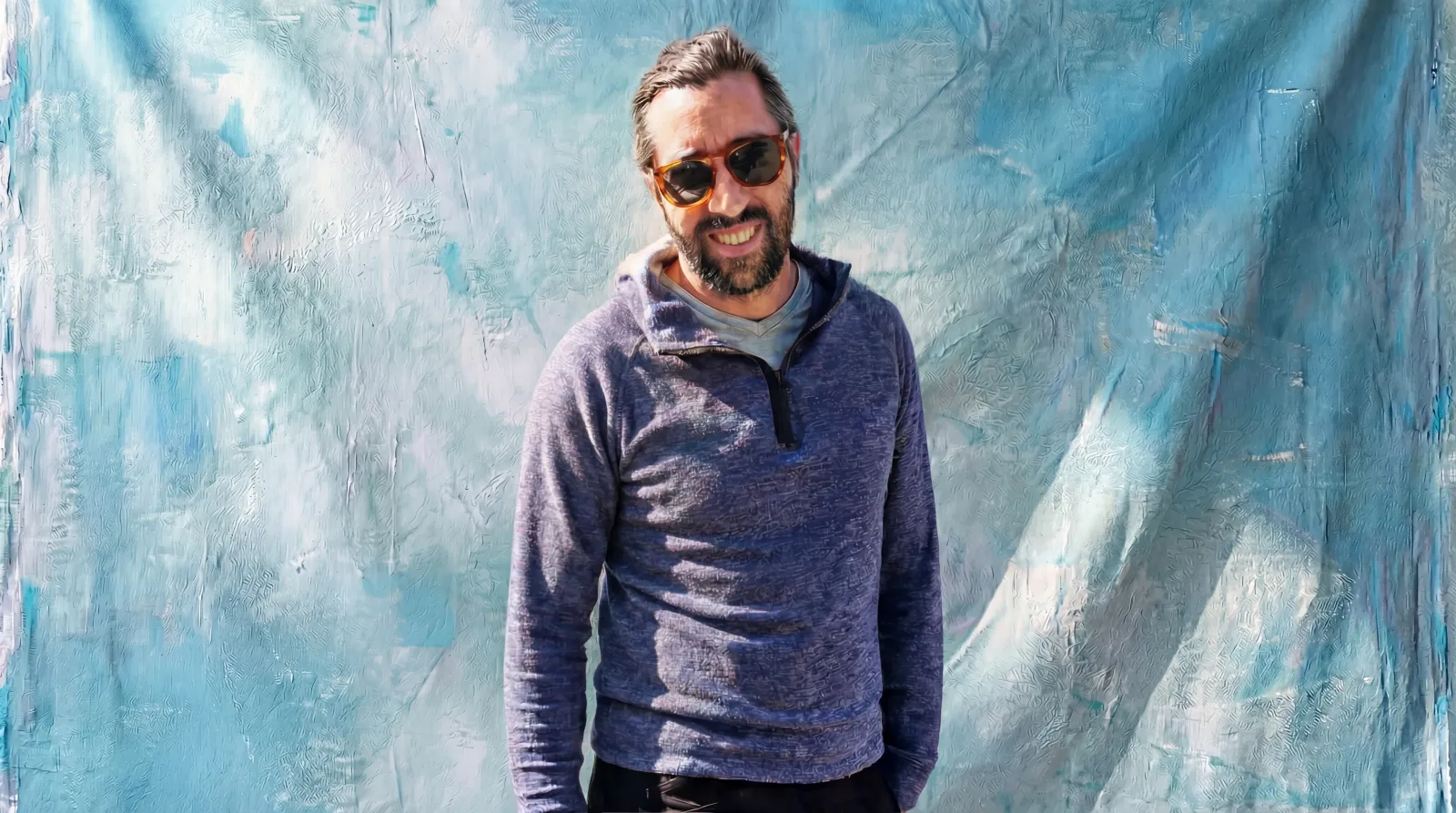Hans Sundquist, Blu Frame’s UI/UX Head of Design, smiling in sunglasses and a blue quarter-zip in front of an artistic turquoise backdrop.