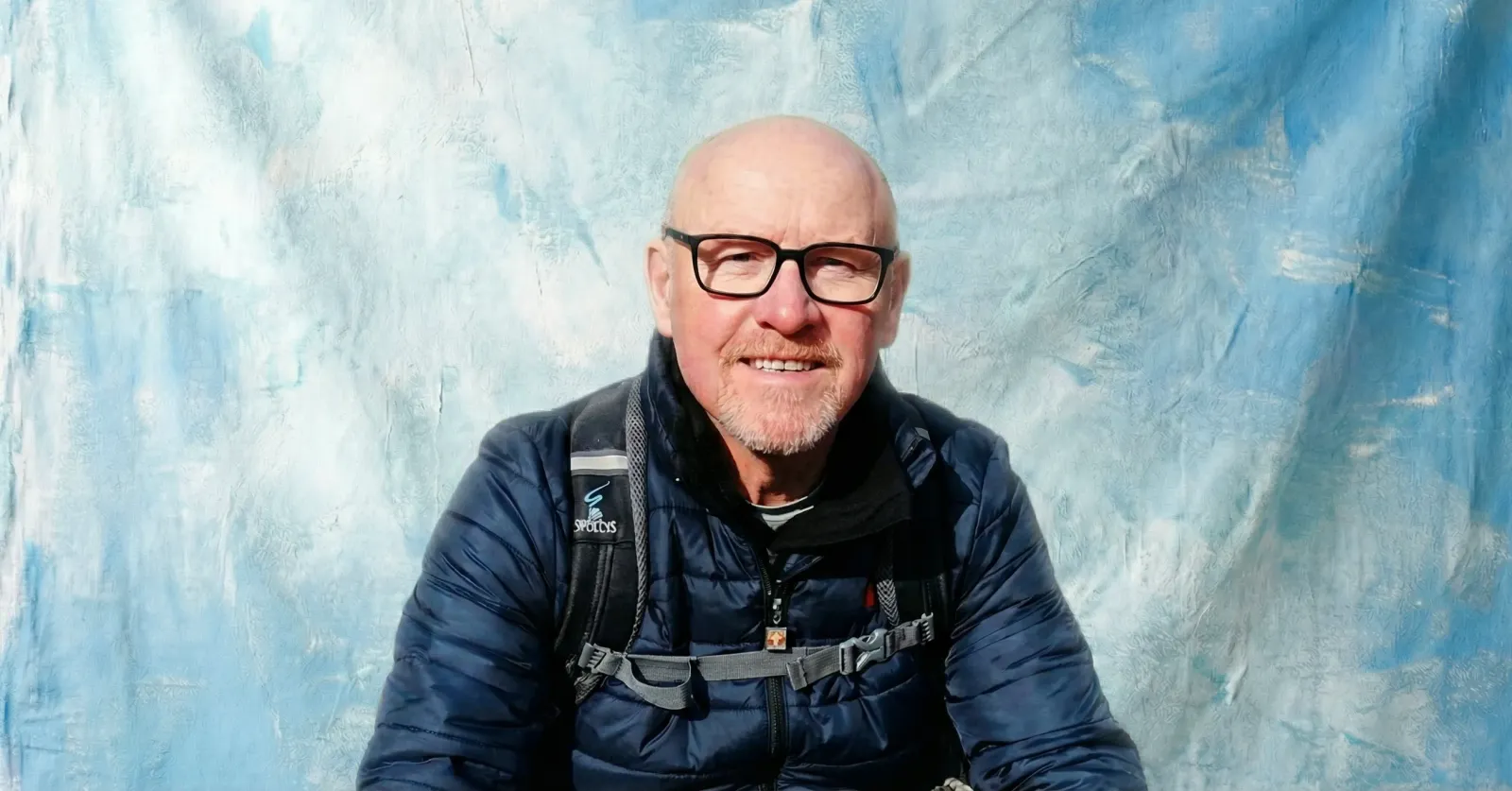 Rob Mansell, Business Development Partner at Blu Frame, smiling in glasses and a navy jacket with a backpack, against a textured blue background.